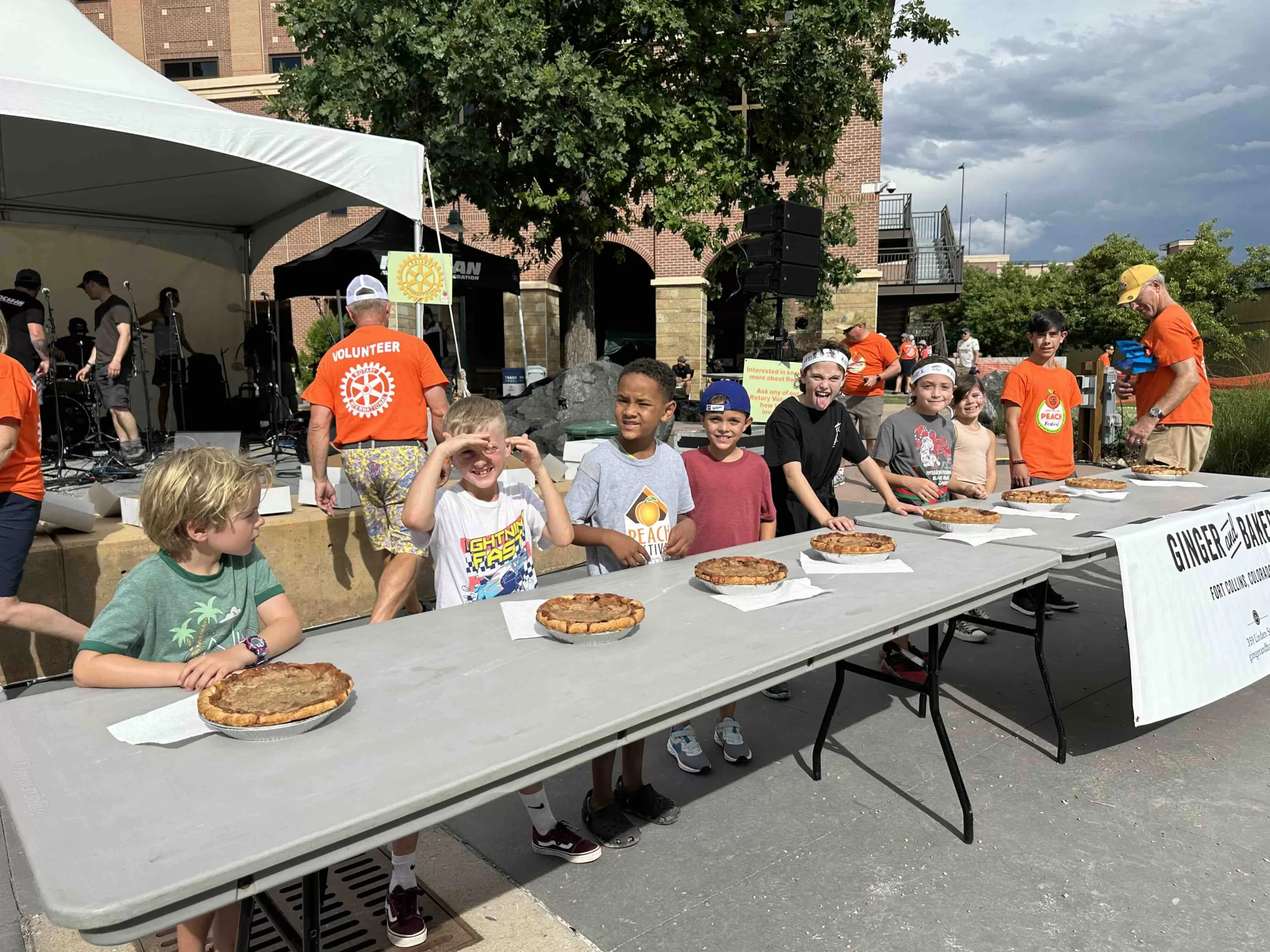 Eight children standing at a folding table, in front of pies, for the pie eating contest.